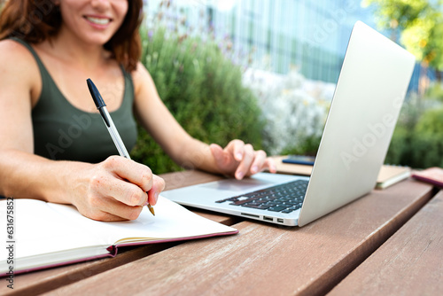 Close up of female college student handwriting on notebook with pen. Young woman using laptop outdoors.