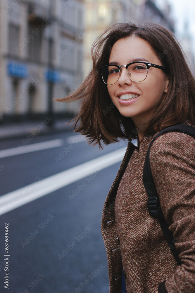 Fototapeta premium A beautiful girl with glasses crosses the road, smiles and looks into the camera. The photo is like a poster, it has a copy space for text