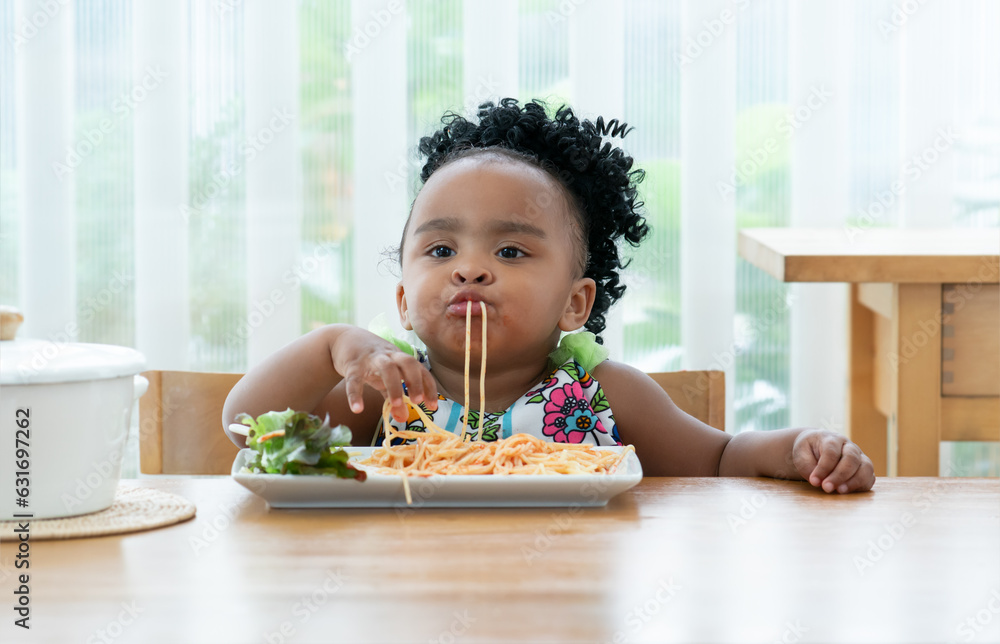 Portrait of little African toddler girl eating spaghetti tomato sauce and vegetables with hand in kitchen at home. Happy messy preschool child eating fresh cooked healthy meal with yummy face Portrait of little African toddler girl eating spaghetti tomato sauce and vegetables with hand in kitchen at home. Happy messy preschool child eating fresh cooked healthy meal with yummy face