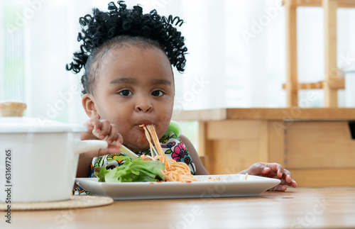 Portrait of little African hungry toddler girl eating spaghetti and vegetables with fork in kitchen at home. Happy messy preschool child looking at camera, face and mouth full of tomato sauce