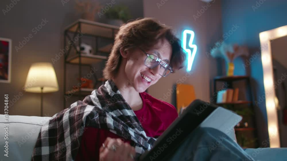 Asian guy in glasses with tablet sits on sofa, online communication. Vivid expressive emotions. Cinematic advertising. Expresses joy, he got good news. Concept of modern virtual lifestyle
