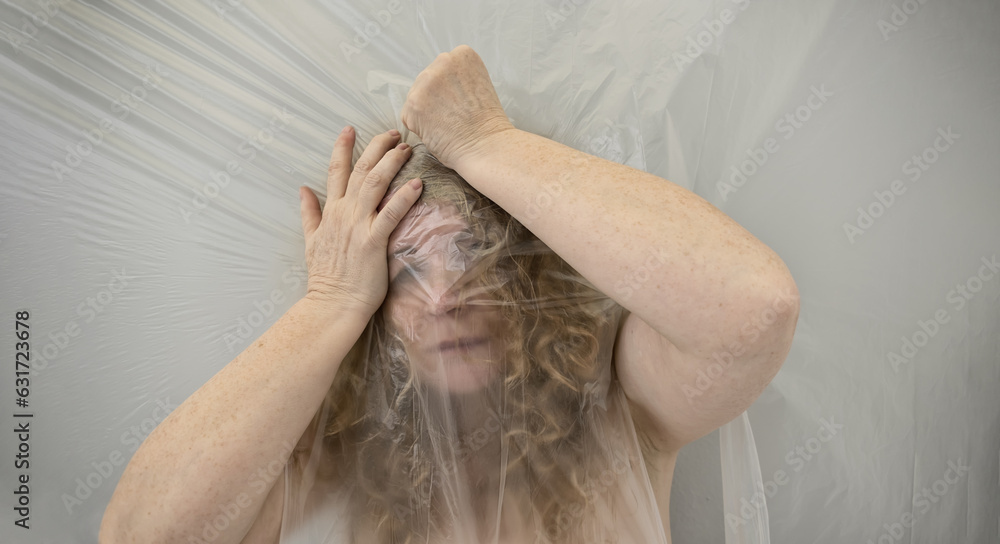 Fototapeta premium blurred image of a harried, stressed, mature woman, grabbing her head from a headache, the position behind transparent plastic film foil represents the altered perception and the dull pain