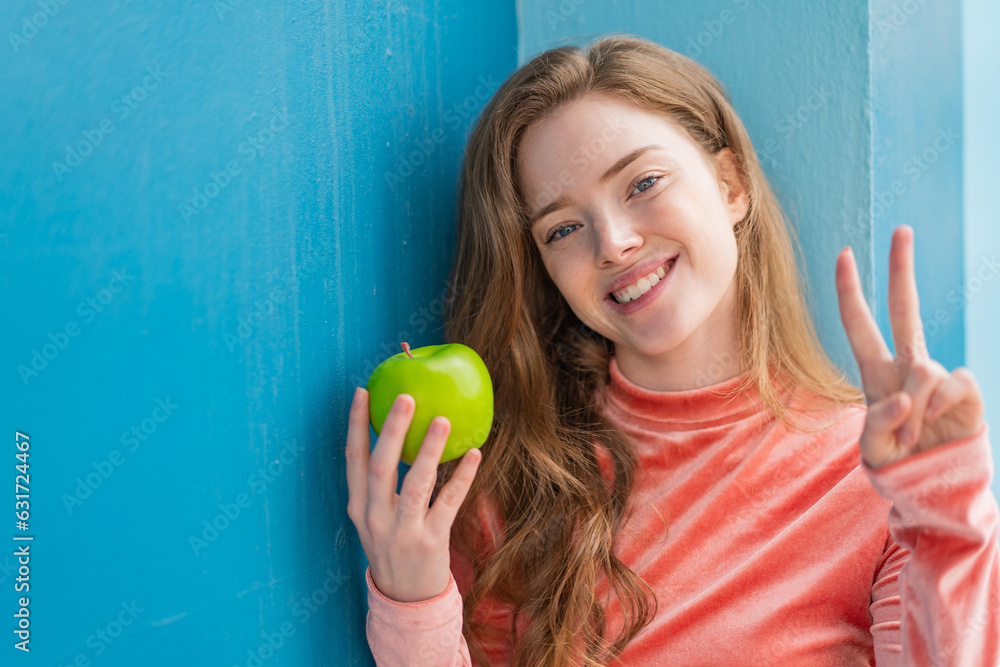 Young redhead woman with an apple at outdoors smiling and showing victory sign Young redhead woman with an apple at outdoors smiling and showing victory sign