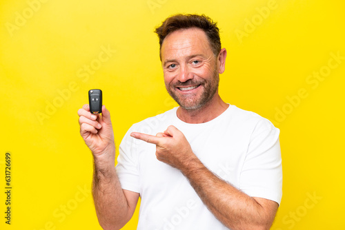 Middle age caucasian man holding car keys isolated on yellow background and pointing it