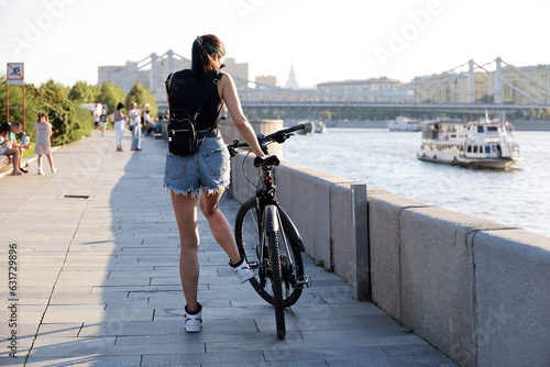 Fototapet Girl in shorts and sunglasses stopped with bicycle on river embankment on city background