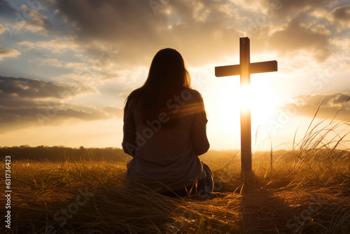 Obraz na plátne Silhouette of a woman sitting on the grass praying in front of a cross at sunset