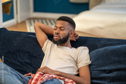Tired pensive black man relaxing on comfortable couch enjoying lazy weekend at modern interior home. . Thoughtful bearded dreaming African American guy sits on sofa rests in studio apartment.
