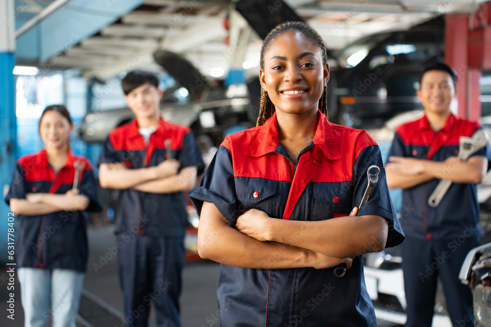 Selective focus of an African female mechanic in uniform, standing to hold a ring spanner with arms folded and smiling at camera with a blurred mechanic team standing in the background in the garage. Selective focus of an African female mechanic in uniform, standing to hold a ring spanner with arms folded and smiling at camera with a blurred mechanic team standing in the background in the garage.