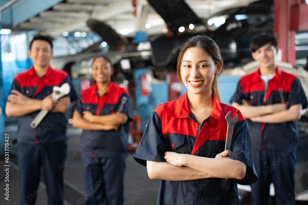 Obraz premium Selective focus of a pretty Asian female mechanic in uniform, standing to hold a wrench with arms folded and smiling at camera, with a blurred mechanic team standing in the background in the garage. 