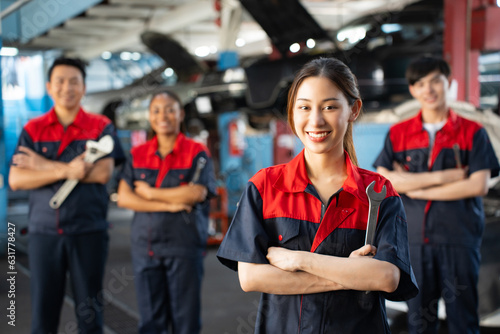 Selective focus of a pretty Asian female mechanic in uniform, standing to hold a wrench with arms folded and smiling at camera, with a blurred mechanic team standing in the background in the garage. 