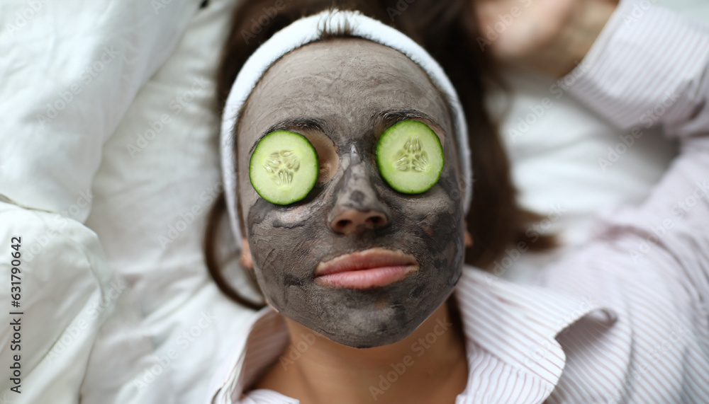 Φωτογραφία Close-up of young beautiful female applying facial clay mask and slices of fresh cucumber on eyes
