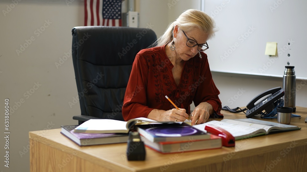 Happy woman teacher sitting at desk in empty school classroom desk grading papers working on assignments. US American flag in the background. Happy woman teacher sitting at desk in empty school classroom desk grading papers working on assignments. US American flag in the background.
