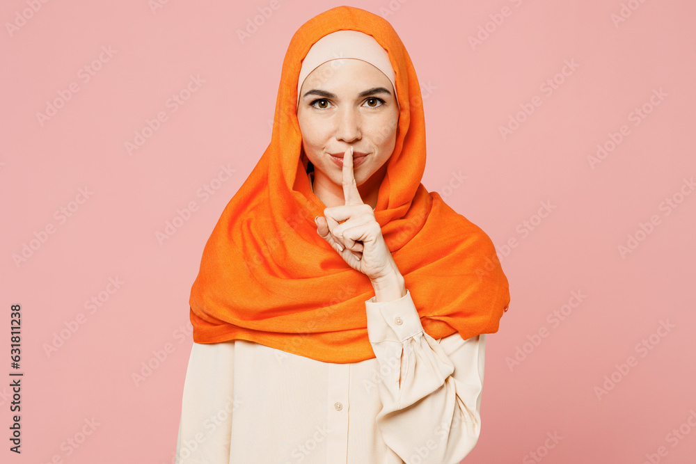 Fototapeta premium Young arabian asian muslim woman wear orange abaya hijab say hush be quiet with finger on lips shhh gesture isolated on plain light pink background studio. Uae middle eastern islam religious concept.