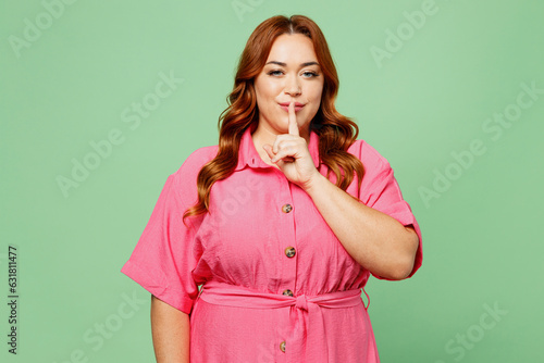 Young secret chubby overweight redhead woman wear casual clothes pink dress say hush be quiet with finger on lips shhh gesture isolated on plain pastel light green color background. Lifesyle concept. photo