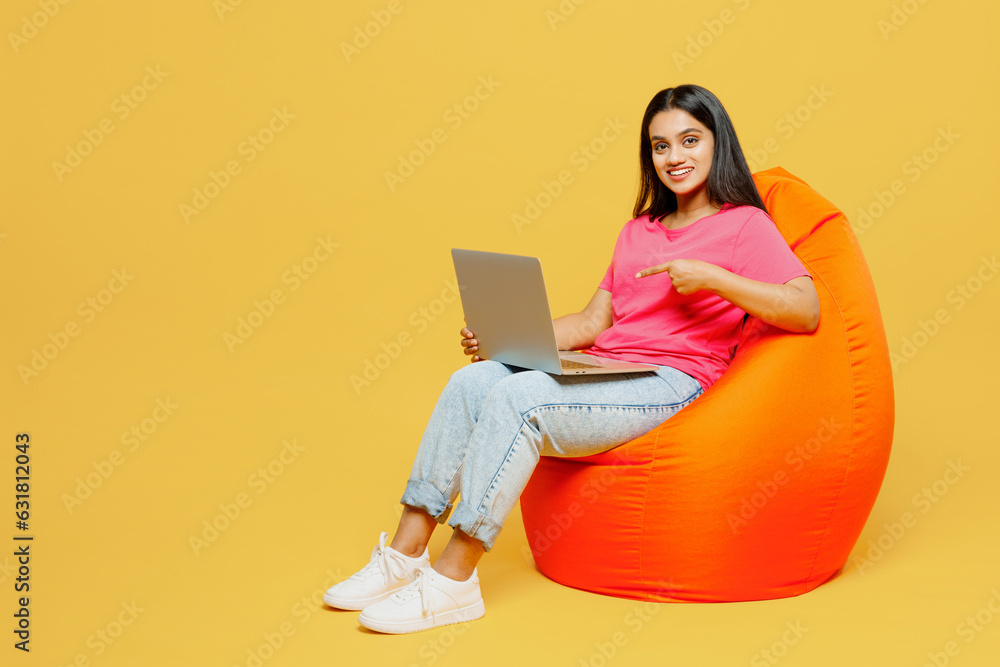 Full body young IT Indian woman wearing pink t-shirt casual clothes sit in bag chair hold use work point on laptop pc computer isolated on plain yellow background studio portrait. Lifestyle concept. Full body young IT Indian woman wearing pink t-shirt casual clothes sit in bag chair hold use work point on laptop pc computer isolated on plain yellow background studio portrait. Lifestyle concept.