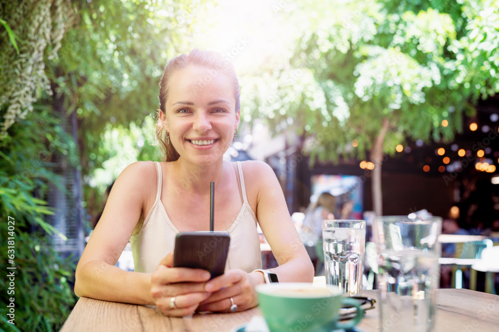 Young adult brunette woman sitting at the table in sidewalk cafe holding smart phone in hand, looking at camera, smiling. Young adult brunette woman sitting at the table in sidewalk cafe holding smart phone in hand, looking at camera, smiling.
