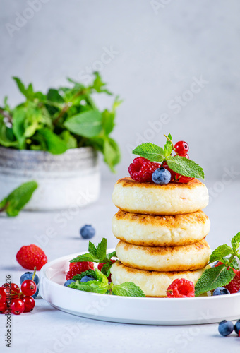 Thick pancakes with cottage cheese with blueberries, raspberries and red currants, decorated with mint on white plate, delicious breakfast. Gray table background