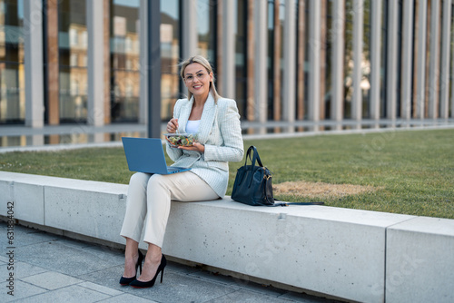 Caucasian young female entrepreneur sitting on a break eating salad. Beautiful business woman looking at camera with a laptop while holding takeaway food.