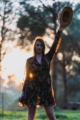 Young woman in a dress and a hat standing in a grassy meadow at the golden hour