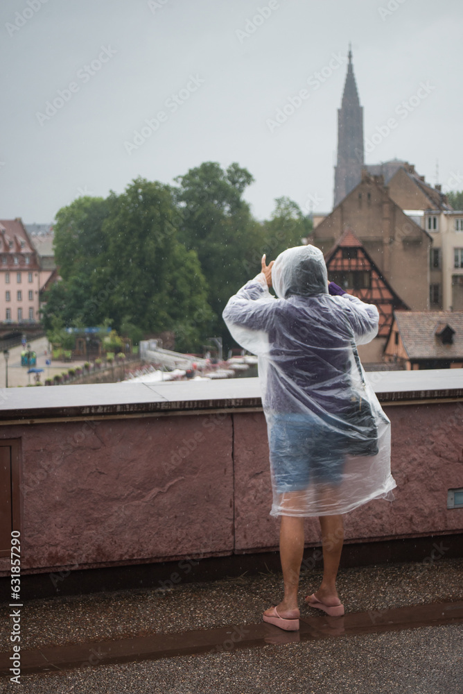 Portrait on back view of woman wearing a transparent rain coat taking a photo with her smartphone in the street
