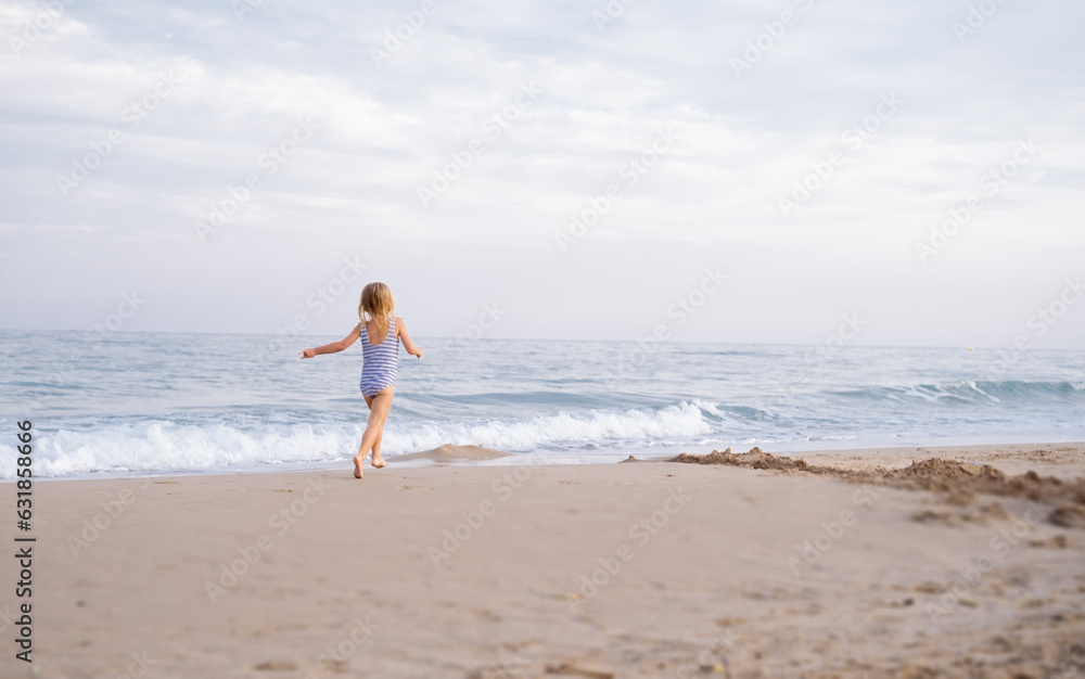 Little girl with long hair running away along sea on sandy beach at Mediterranean seaside in Spain. Carefree childhood, freedom, happiness concept.Back view. Little girl with long hair running away along sea on sandy beach at Mediterranean seaside in Spain. Carefree childhood, freedom, happiness concept.Back view.