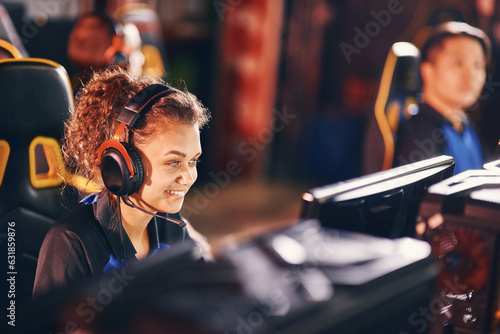 Smiling mixed race girl, female gamer wearing headphones playing online video games while sitting in gaming club or internet cafe