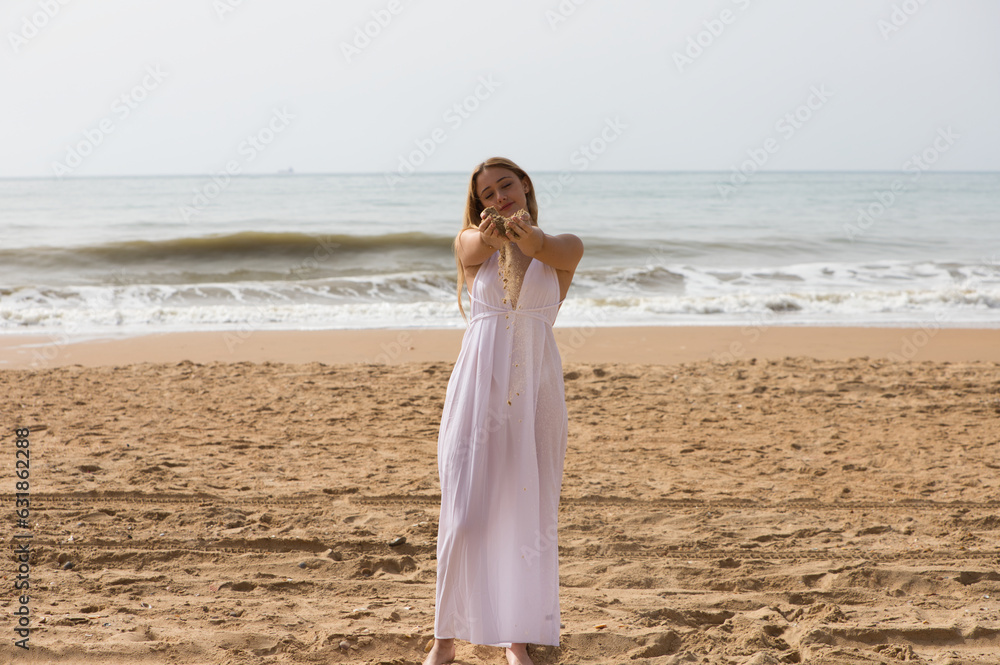 Pretty young blonde woman in white dress spills sand from between her hands as she holds her hands out in front of her. The woman is happy and having fun. In the background the blue sea.