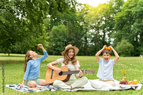 Cute blond teenager girl in a straw hat plays the guitar sitting on the grass in the city park. Younger brother and sister sit on the blanket and play with food, have fun. Three children at a picnic
