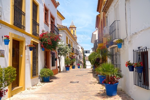 Fototapeta Naklejka Na Ścianę i Meble -  narrow beautiful street in the old town of Estepona with lots of flowers and a view to the church tower of Parroquia Nuestra Señora De Los Remedios, Málaga, Andalusia, Spain