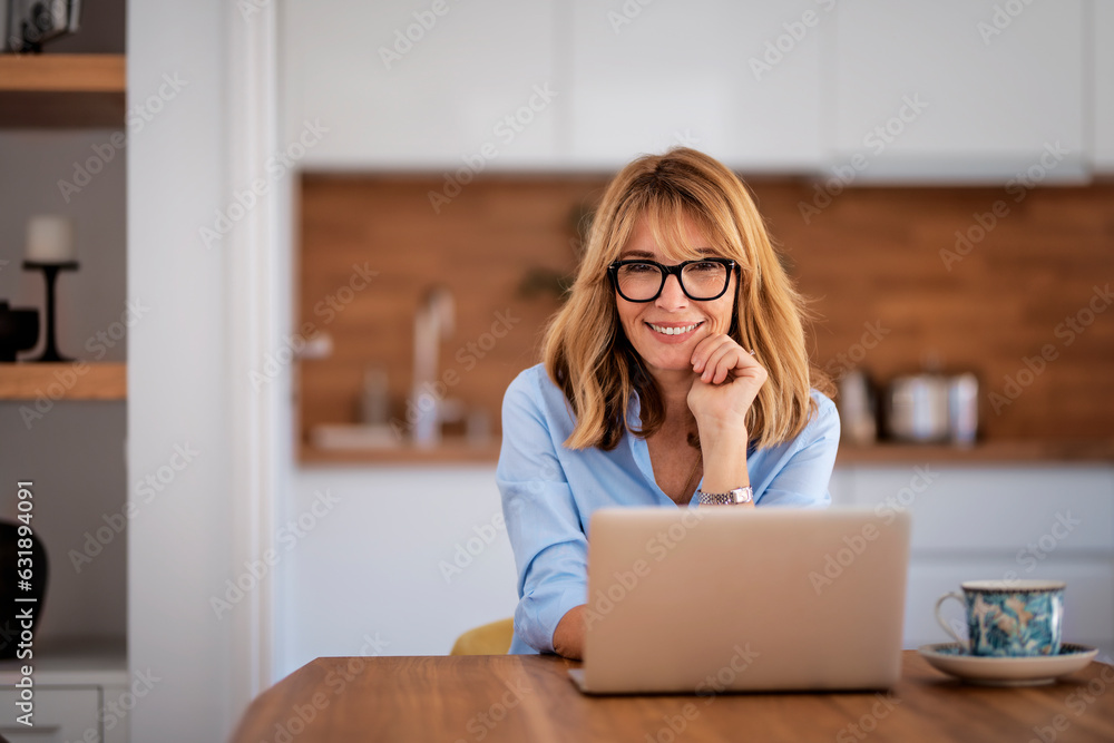 Laughing mid aged woman sitting in her dining room and using laptop for work Laughing mid aged woman sitting in her dining room and using laptop for work