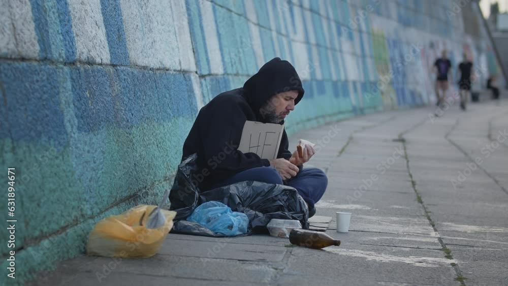 homeless poor young man sitting on the ground in the city with a Help table while asking for money to survive. Eating old unhealthy food homeless poor young man sitting on the ground in the city with a Help table while asking for money to survive. Eating old unhealthy food