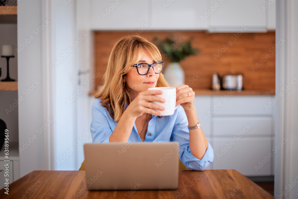 Smiling mid aged woman drinking tea and using laptop at home Smiling mid aged woman drinking tea and using laptop at home