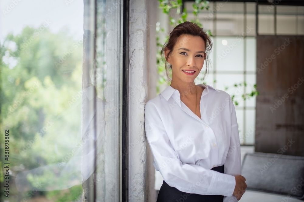 Portait of an attractive mid aged woman wearing white shirt and black pants and standing by the window
