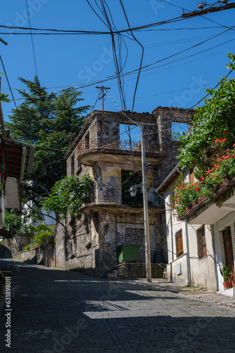 Abandoned ruined stone building on a cobblestone street in Gjirokaster, Albania, with power lines and green trees in summer sunlight.

