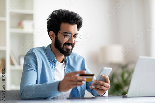 Mobile Banking. Smiling Indian Man Using Smartphone And Credit Card In Office