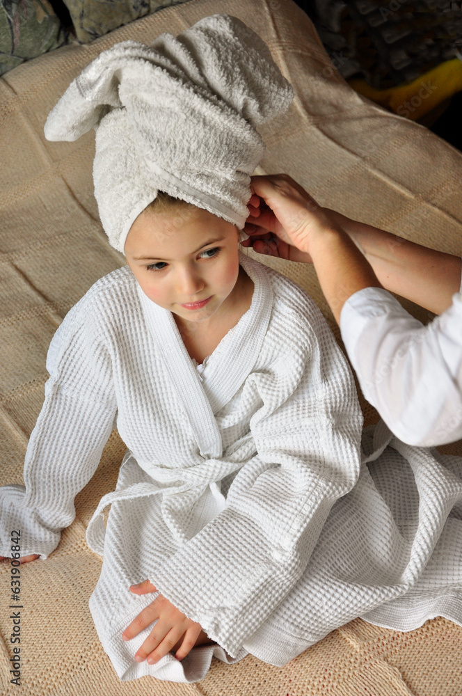 A girl in a bathrobe sits on a bed in the bedroom. Mom wraps the girl's hair in a towel. A girl in a bathrobe sits on a bed in the bedroom. Mom wraps the girl's hair in a towel.