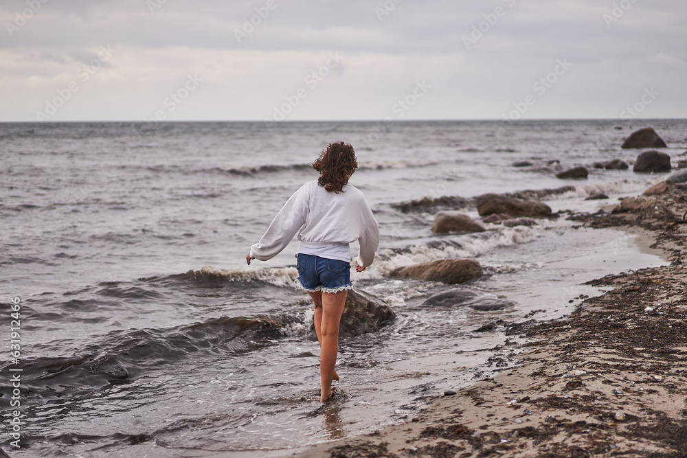 Teenage girl walking alone along water line on the sand and stone beach of North sea on the coast of Denmark during her summer vacation in scandinavian countries and enjoying water, wind and fresh air