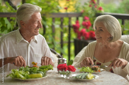 Happy elderly man and woman in casual outfits sitting at kitchen table  having healthy breakfast together at home  eating organic food  drinking fresh juice and chatting  copy space