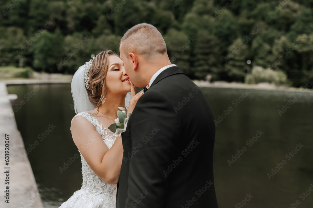 A brunette bride in a long dress and a groom in a classic suit stand embracing on a bridge near a lake with a castle in the background.