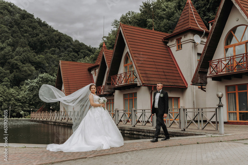 A brunette bride in a long dress and a groom in a classic suit are standing on a bridge near a lake against the background of a castle. A veil is thrown in the air