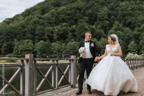 A brunette bride in a long dress and a groom in a classic suit are walking on a bridge near a lake against the background of a castle. a walk in nature. Wedding day