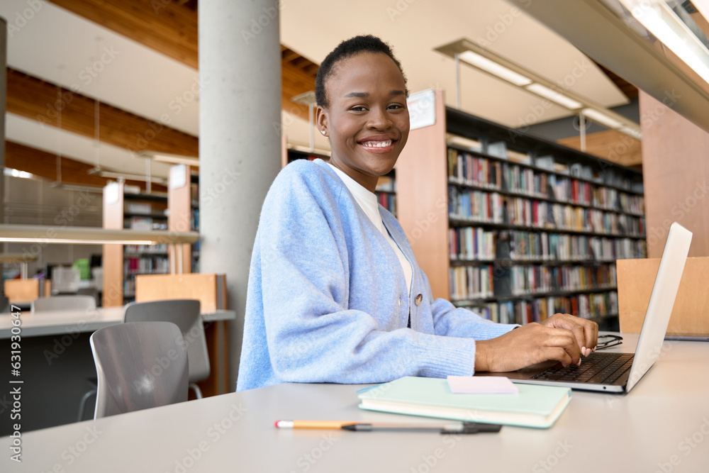 Happy African Black girl student studying in university library using laptop, school girl sitting at desk in college campus elearning, studying, hybrid learning online, looking at camera, portrait. Happy African Black girl student studying in university library using laptop, school girl sitting at desk in college campus elearning, studying, hybrid learning online, looking at camera, portrait.