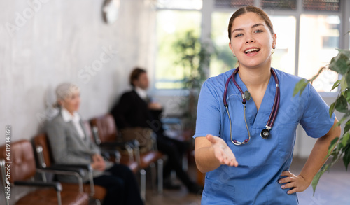 Cheerful friendly young female doctor in blue uniform making welcome gesture, politely inviting patients in medical office