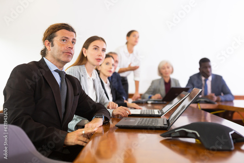 Mid aged white dark-haired male manager attending business meeting in conference room and interestedly watching colleague's presentation together with coworkers sitting in row behind him