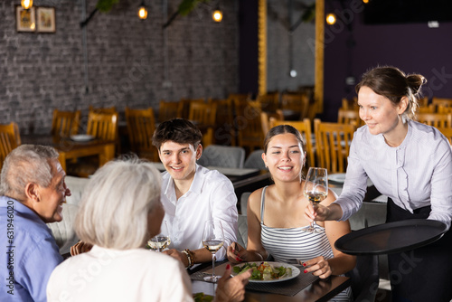 Adult woman waiter serving glass of wine to elderly couple and young couple in restaurant