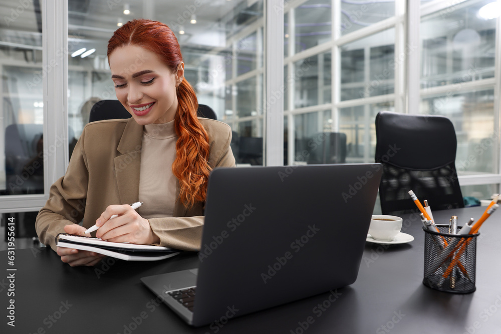 Happy woman taking notes while working with laptop at black desk in office Happy woman taking notes while working with laptop at black desk in office