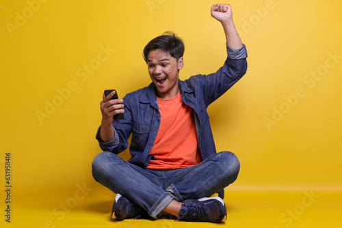 Schilderij op canvas Full length of delighted Asian man using cellphone and raising fist, show victory gesture while sitting on floor with crossed legs isolated over yellow studio background