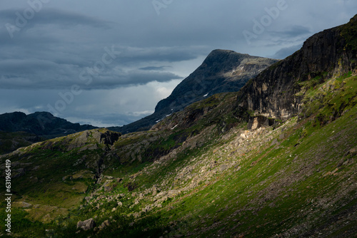 Sunlit green mountain slopes on the way up to Rabothytta, Okstindan, Hemnes, Helgeland, Northern Norway. Green mountains of Nordnorge. Sunny patches on dark mountains. Cloudy dramatic skies and light. photo