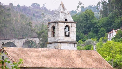 Up close, the bell at church of São Tiago de Valadares in Baião stands prominent. Behind, a bridge adds to the quaint charm of the scene photo