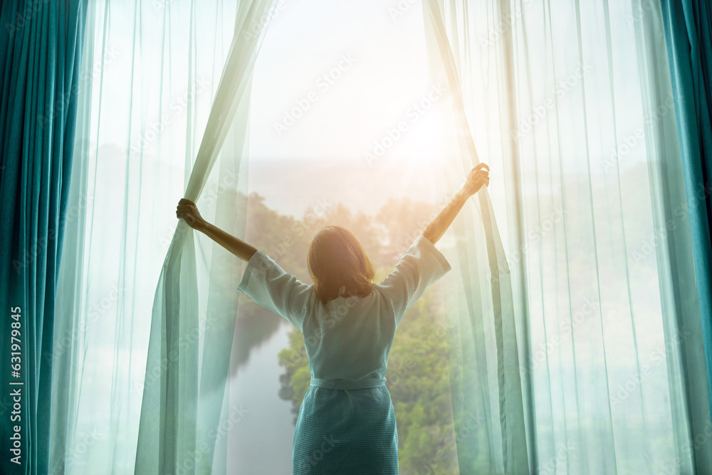 Fototapeta premium The woman was at the window in the bedroom. She opened the curtains on the window. In the morning and she looks at the view of mountains and trees at sunrise.